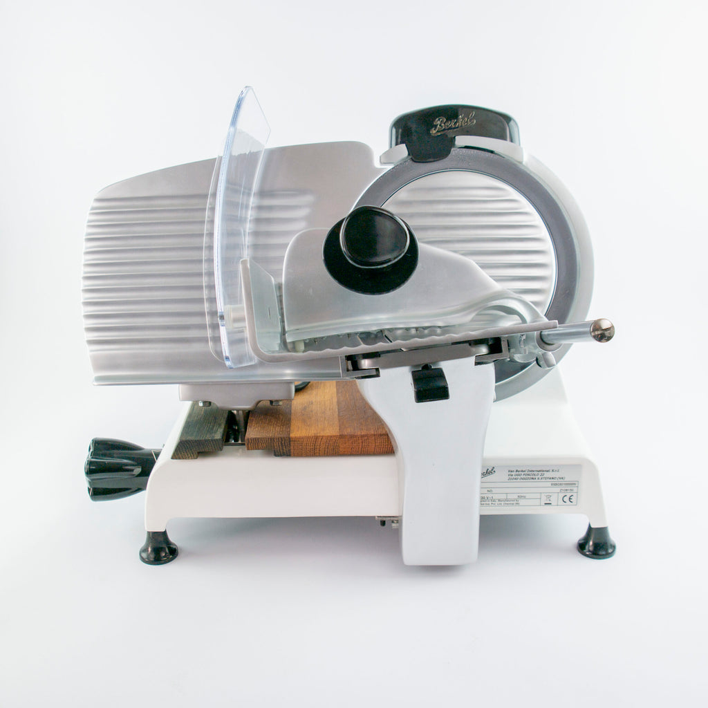 Ein silber-weißer elektrischer Fleischschneider mit gezahnter Klinge, Schutzvorrichtung, Holzschieber (Einlegebrett aus Fassholz) und einstellbarem Dickenregler auf einer weißen Oberfläche. Die Berkel Red Line 250 Weiss Original Profi-Aufschnittmaschine verfügt über einen Klingendurchmesser von 25 cm und ist für das präzise Schneiden von Fleisch und Käse konzipiert.