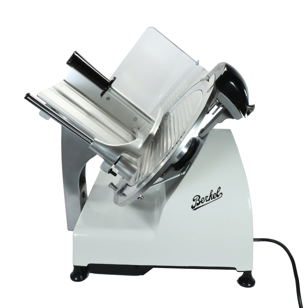 Die Berkel Red Line Weiß, 300 mm Original Profi-Aufschnittmaschine + Weinfass Einlegebrett ist eine elegante, weiße Aufschnittmaschine mit runder Klinge und transparentem Schutzgitter. Die elektrische Aufschnittmaschine wird mit einem angebrachten Lebensmittelschlitten zum Schneiden von Fleisch geliefert und steht elegant auf einem schlichten weißen Hintergrund.