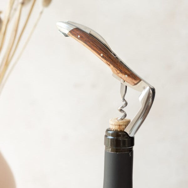 Ein Kellnermesser-Sommelier-Korkenzieher von Forge De Laguiole mit handgefertigtem Thuja-Griff ist anmutig in den Korken einer Weinflasche eingesetzt, vor einem verschwommenen Hintergrund aus hellen Trockenblumen.