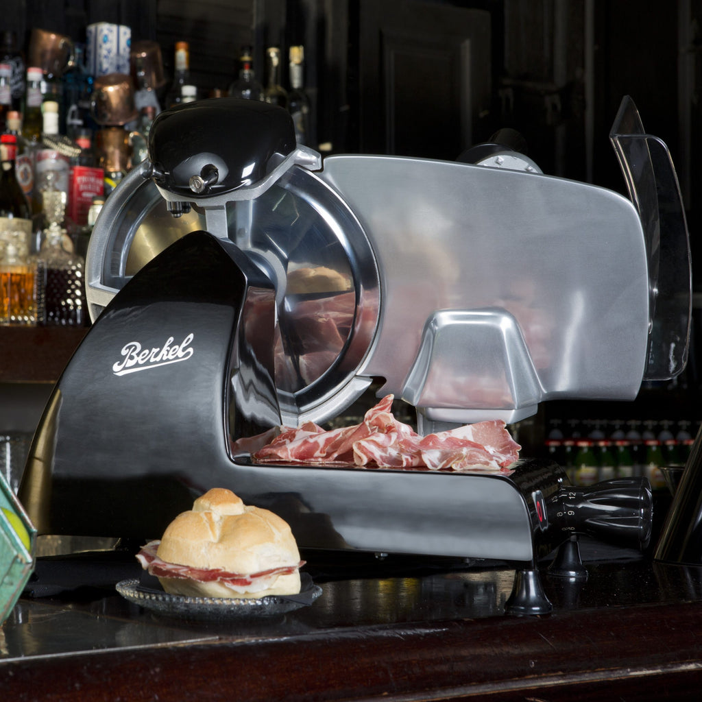 Eine Berkel Red Line 250 Schwarz Profi-Aufschnittmaschine mit 25 cm Schneideblatt schneidet Aufschnitt neben einem belegten Sandwich auf eine Holztheke, im Hintergrund sind Flaschen zu sehen.