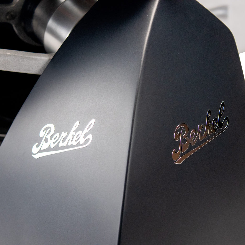 Eine Nahaufnahme des Berkel RedLine 250 MATT in Schwarz mit Fassholzbrett zeigt sein schlankes, modernes Design. Der Name „Berkel“ ist stilvoll zweimal in metallischer Schrift eingraviert und unterstreicht seine glänzende Attraktivität und den prestigeträchtigen Markennamen.