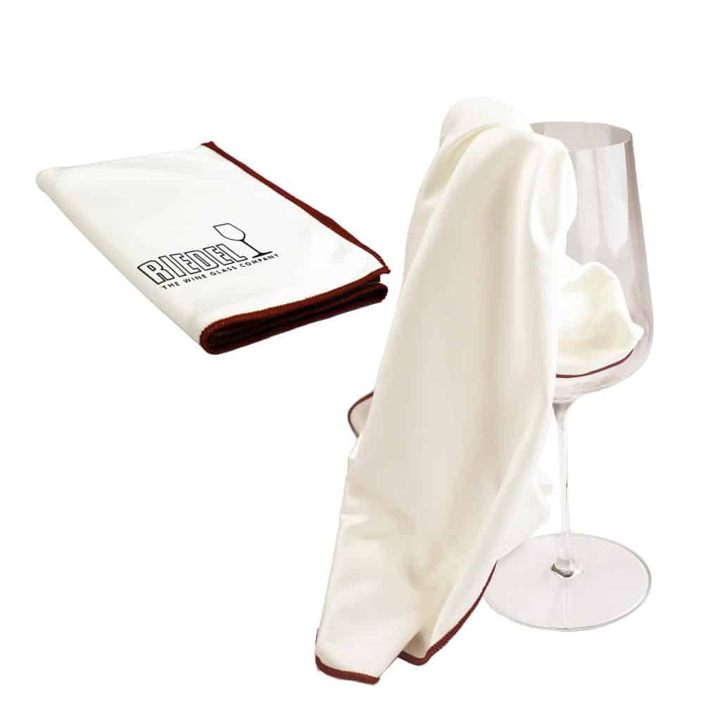 Ein weißes Riedel Microfasertuch mit braunen Kanten ist gefaltet abgebildet und trägt den Markennamen „Riedel“ und ein Weinglas-Logo. Ein weiteres Tuch liegt über einem leeren Weinglas und demonstriert seine Verwendung zum Polieren.
