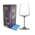 Ein Weißweinglas Riedel Winewings Chardonnay Glas mit Poliertuch in Geschenkverpackung steht neben seiner Verpackung und einem gefalteten blauen Tuch. Die Geschenkverpackung ist größtenteils schwarz mit violetten Akzenten und zeigt ein Bild des Weinglases. Der Text auf der Verpackung gibt die Marke und das Modell an, während das Glas ein schlankes, modernes Design aufweist.