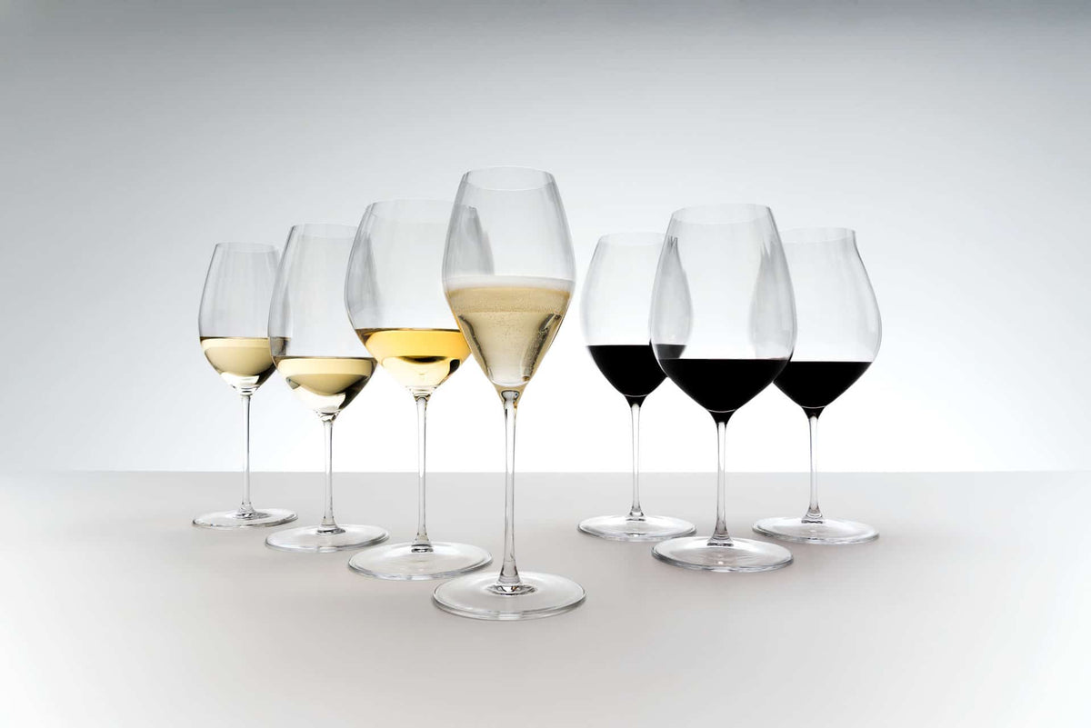 Eine Präsentation von sieben Weißweingläsern Riedel Performance Chardonnay mit Poliertuch, die jeweils mit unterschiedlichen Weinsorten und -mengen gefüllt sind. Die Getränke variieren in der Farbe von weiß bis rot, wobei einige Gläser teilweise gefüllt und andere leer erscheinen. Die Riedel Performance-Gläser sind versetzt vor einem hellen Hintergrund angeordnet.