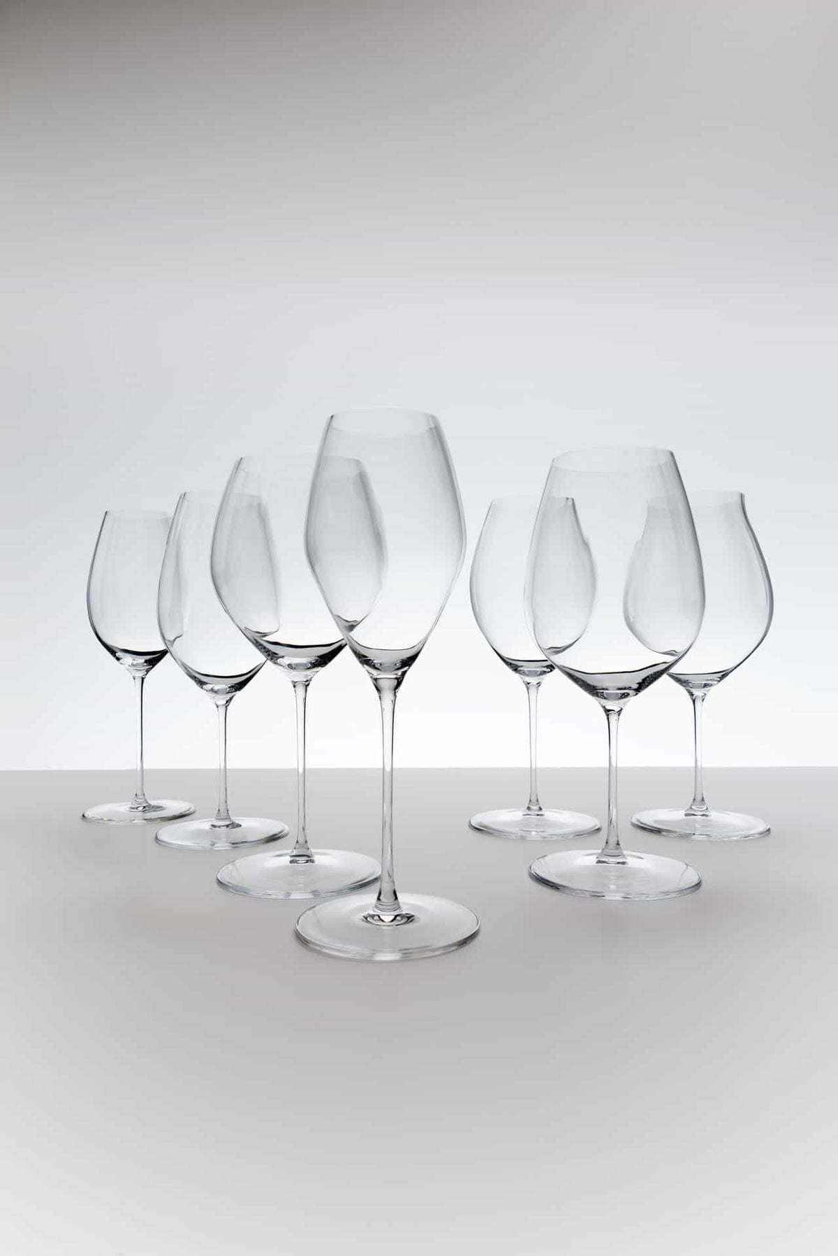 Sechs leere Weißweingläser Riedel Performance Chardonnay mit Poliertuch, ordentlich und leicht versetzt angeordnet auf einer hellen Oberfläche vor einem Hintergrund mit Farbverlauf von Weiß nach Grau. Die eleganten Gläser sind hoch und haben schlanke Stiele, die subtile Reflexe und Schatten auf die Oberfläche werfen.