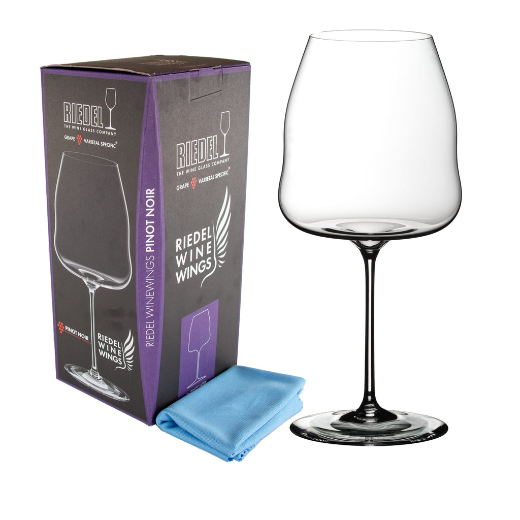Ein Rotweinglas Riedel Winewings Pinot Noir mit Poliertuch in Geschenkverpackung steht neben seiner Schachtel, die größtenteils schwarz und lila ist und einen beschreibenden Text und ein Bild des Glases darauf trägt. Vor der Schachtel liegt ein hellblaues Reinigungstuch.