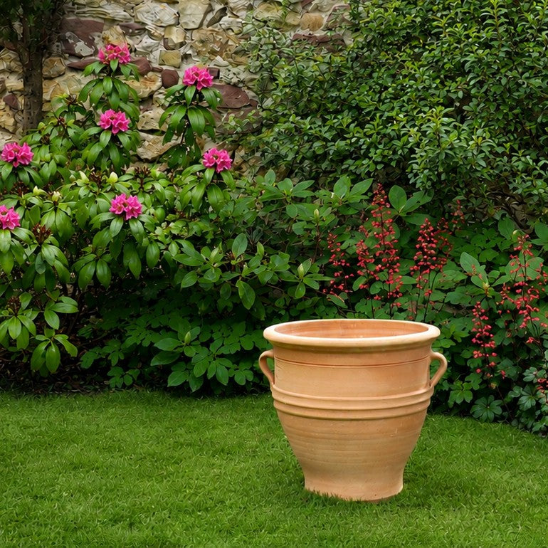 Das Terrakotta-Pflanzgefäß "Rosa", H=40 cm steht auf einer grünen Wiese, umgeben von grünen Büschen mit rosa und roten Blüten, mit einer Steinmauer im Hintergrund.