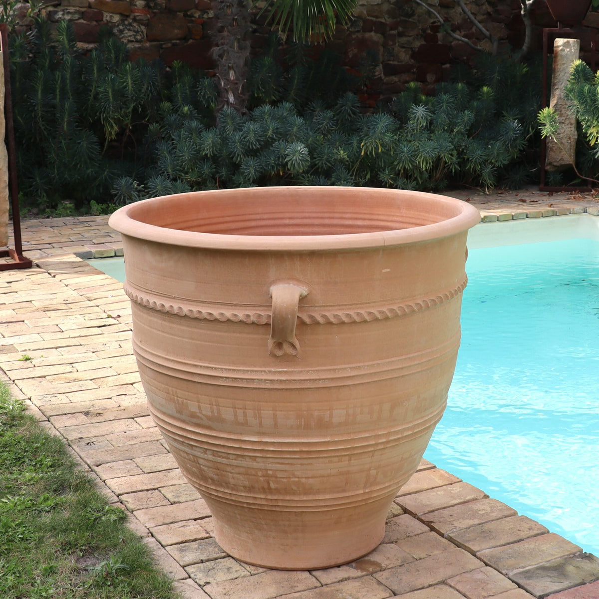 Ein 80 cm hoher Terrakotta-Pflanzkübel „Rosa“ in XXL-Größe steht auf einer gepflasterten Fläche neben einem Swimmingpool. Der große Topf hat Griffe und ist mit horizontalen Rillen und Mustern verziert. Im Hintergrund sind üppiges Grün und ein Teil einer Steinmauer zu sehen.