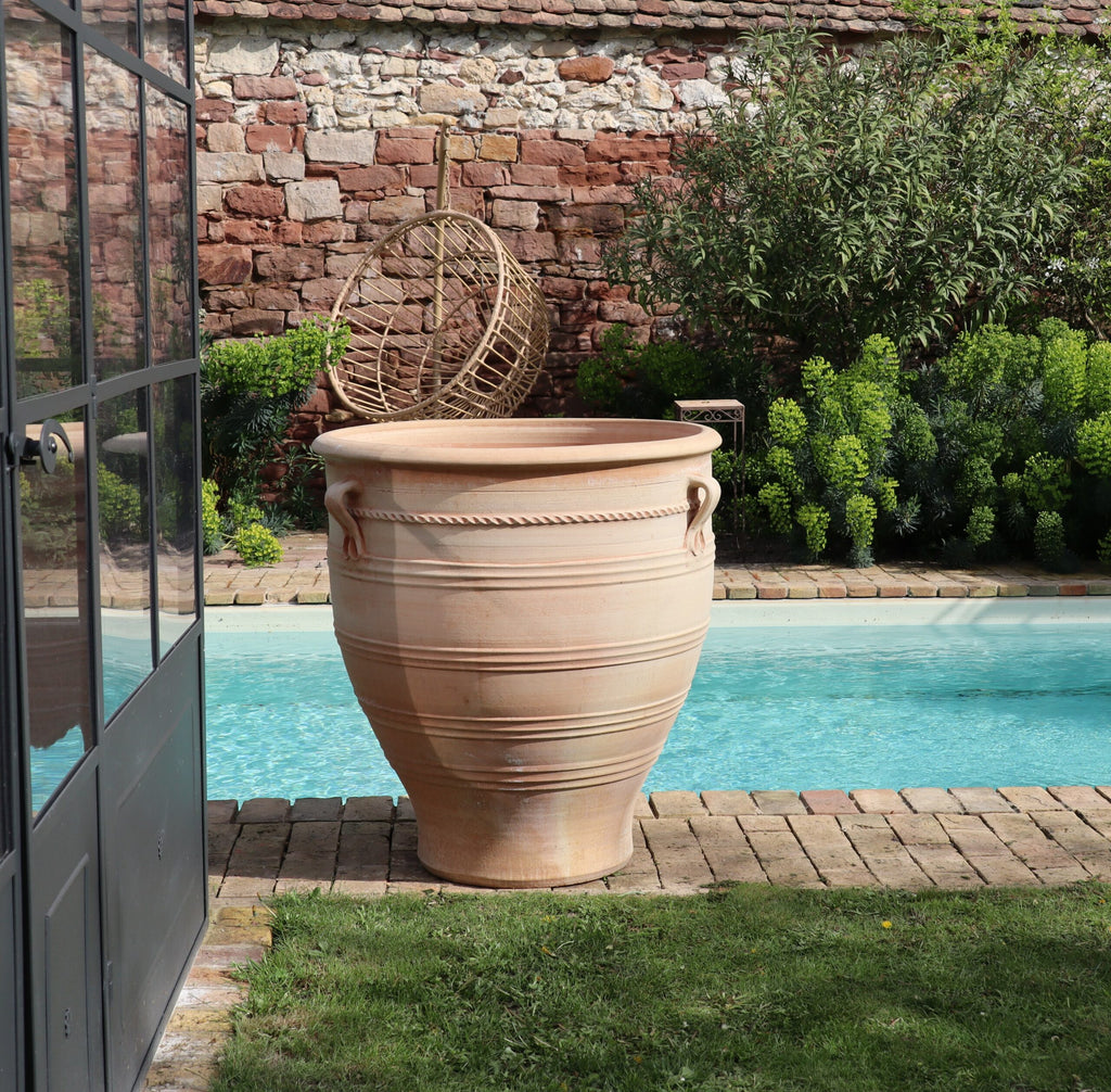 Ein Terrakotta-Pflanzkübel groß, XXL „Rosa“, H=90 cm steht neben einem kleinen Swimmingpool in einem Garten mit Grünflächen und einer Steinmauer im Hintergrund. Ein Korbstuhl lehnt an der Mauer. Der Poolbereich ist mit Ziegeln eingefasst, links sind Glastüren sichtbar, die einen subtilen Hauch von Rosa-Charme verströmen.