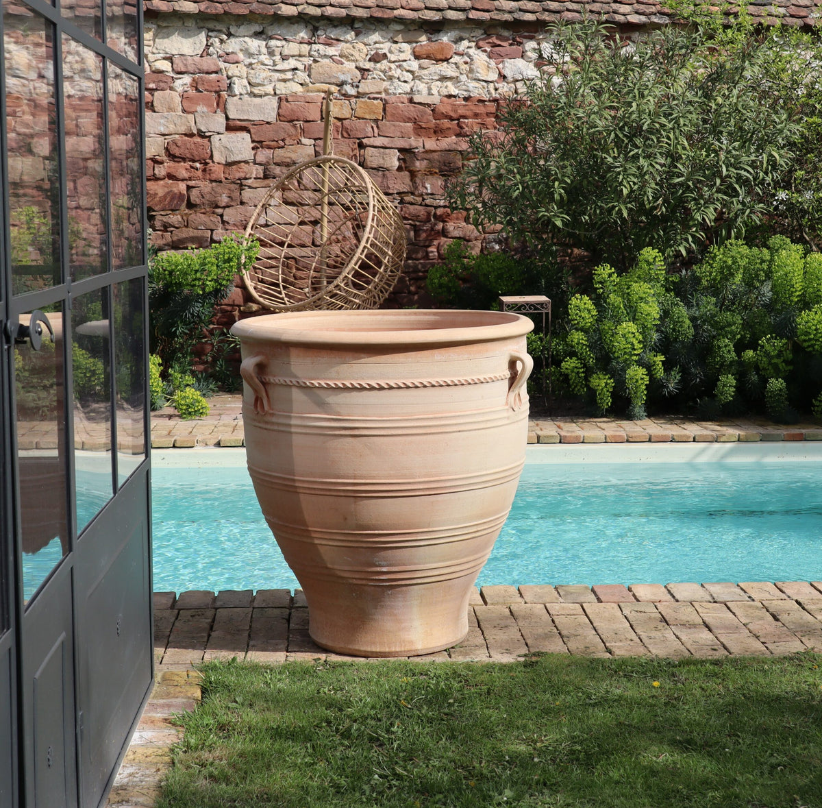Ein Terrakotta-Pflanzkübel groß, XXL „Rosa“, H=90 cm steht neben einem kleinen Swimmingpool in einem Garten mit Grünflächen und einer Steinmauer im Hintergrund. Ein Korbstuhl lehnt an der Mauer. Der Poolbereich ist mit Ziegeln eingefasst, links sind Glastüren sichtbar, die einen subtilen Hauch von Rosa-Charme verströmen.