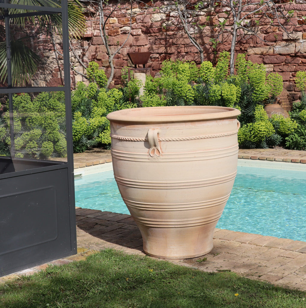 Ein großer, beigefarbener Terrakotta-Pflanzkübel groß, XXL „Rosa“, H=90 cm mit dekorativen Rillen und einem stilisierten Seilmotiv steht neben einem Swimmingpool. Er steht in der Nähe eines Glasgebäudes und vor einem lebendigen Garten mit üppigem, grünem Laub vor einer Backsteinmauer im Hintergrund. Das Poolwasser ist klar und einladend.