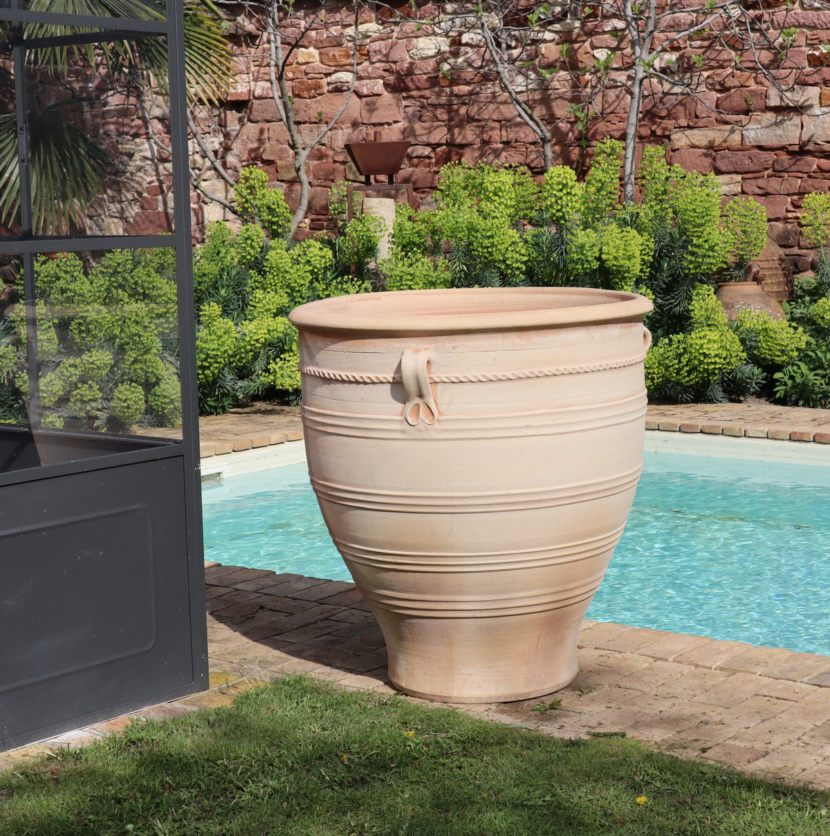 Ein großer, beigefarbener Terrakotta-Pflanzkübel groß, XXL „Rosa“, H=90 cm mit dekorativen Rillen und einem stilisierten Seilmotiv steht neben einem Swimmingpool. Er steht in der Nähe eines Glasgebäudes und vor einem lebendigen Garten mit üppigem, grünem Laub vor einer Backsteinmauer im Hintergrund. Das Poolwasser ist klar und einladend.