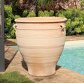 Ein großer, hellbeiger Terrakotta-Pflanzkübel groß, XXL „Rosa“, H=90 cm steht neben einem Pool in einem Gartenbereich im Freien. Die Urne hat zwei kleine Griffe und dekorative Rillen, die horizontal entlang ihrer Oberfläche verlaufen. Im Hintergrund sind üppige grüne Sträucher und eine Backsteinmauer zu sehen.