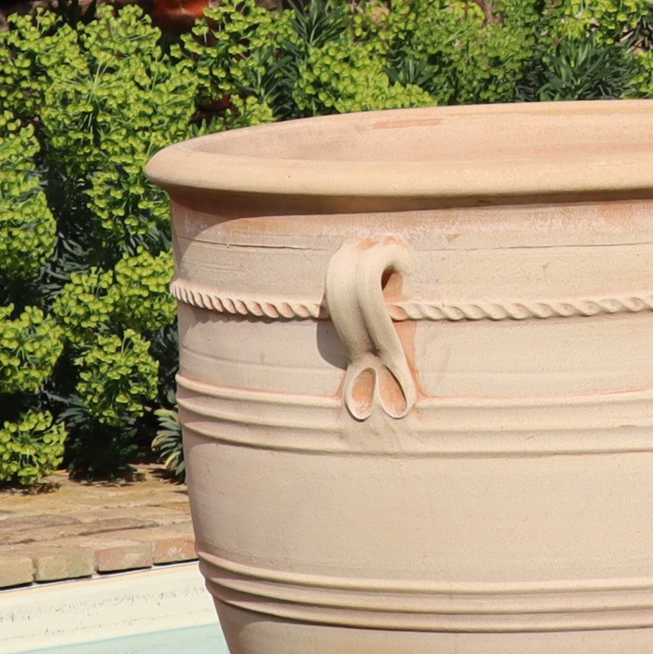 Ein großer, beigefarbener Terrakotta-Pflanzkübel namens „Rosa“ steht im Freien inmitten von grünem Laub und Mauerwerk. Dieses XXL-Stück ist 90 cm hoch und verfügt über dekorative Griffe, komplizierte Muster aus gedrehten Seilen und geformten Schlaufen, was es zu einer herausragenden Ergänzung für jeden Garten macht.