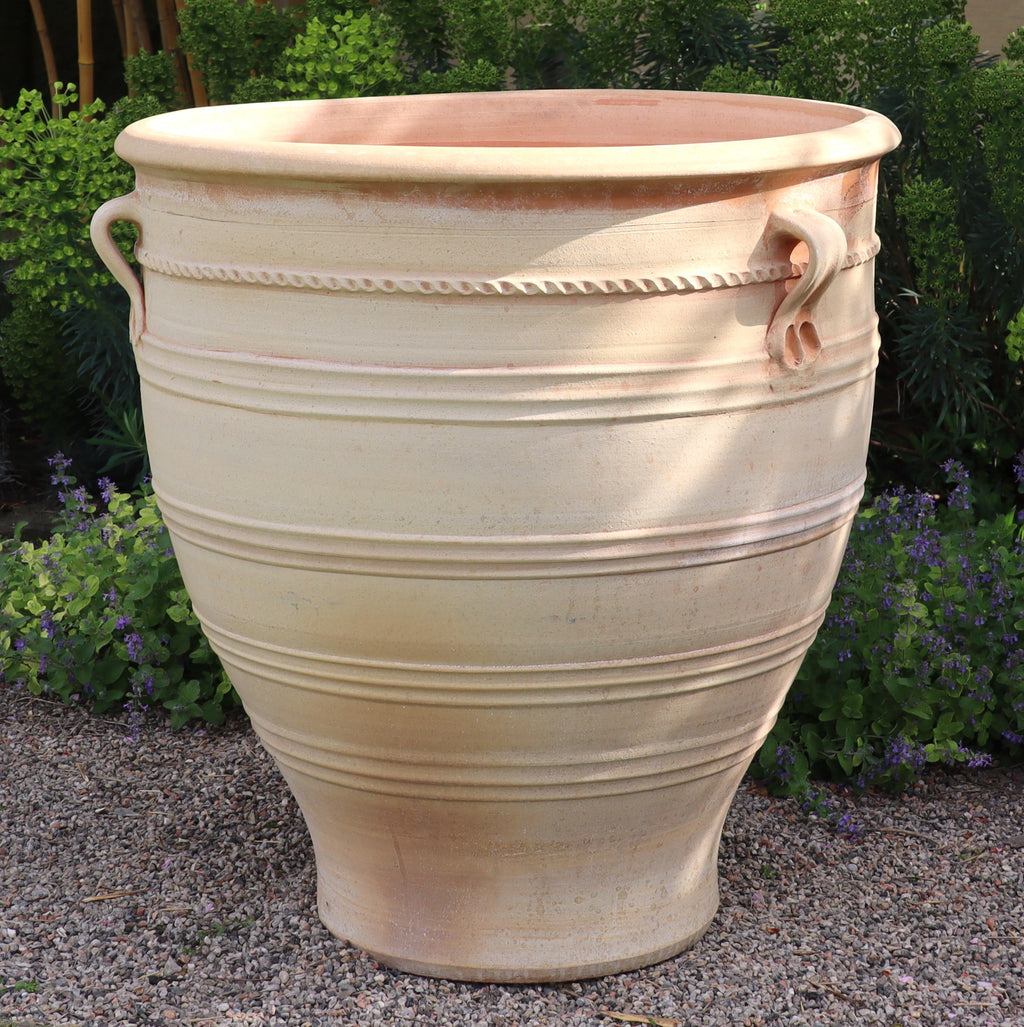 Ein Terrakotta Pflanzkübel groß, XXL „Rosa“, in Beige mit dekorativen Griffen und waagerechten Rillen steht auf einer Kiesfläche. Der 90 cm hohe Topf ist von üppigem Grün und kleinen lila Blüten im Hintergrund umgeben und suggeriert so eine Gartenkulisse.