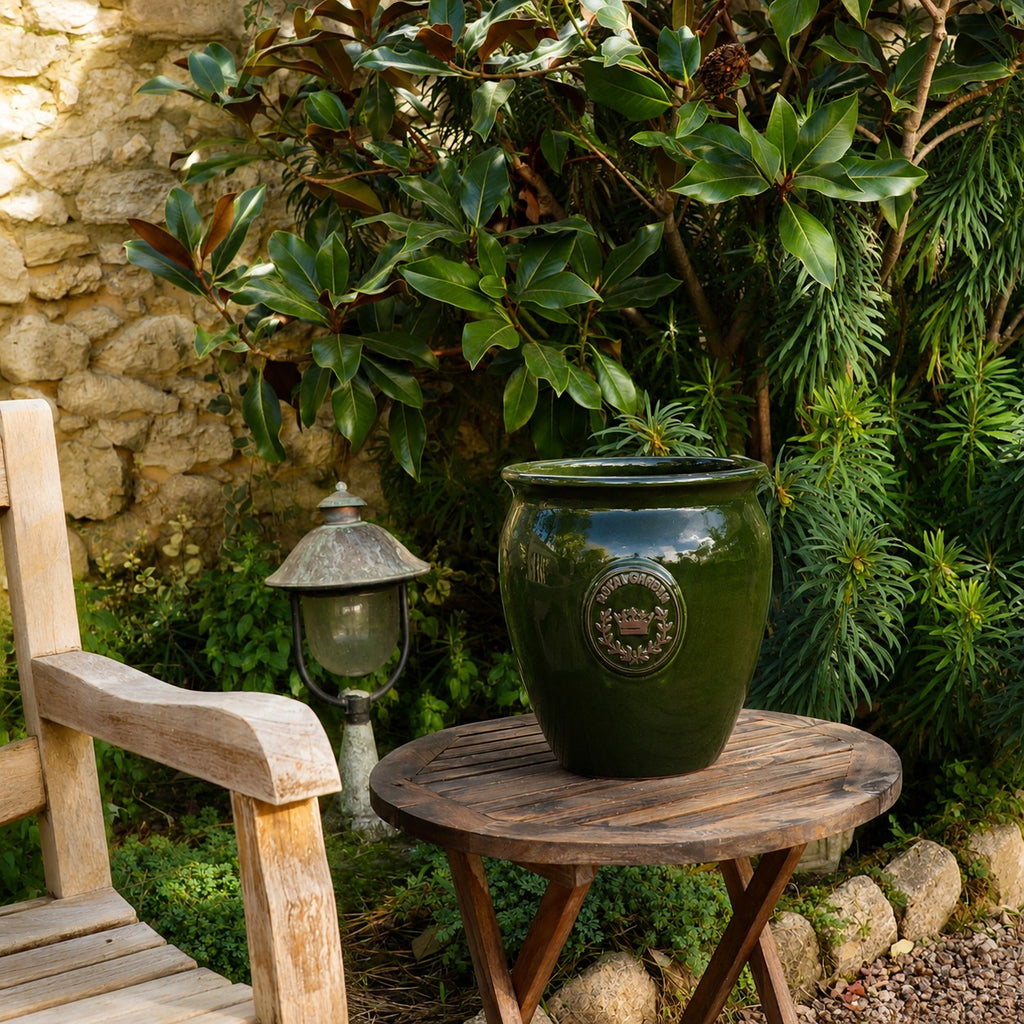 Der grün glasierte Blumentopf „eggpot“ (Höhe: 29–46 cm) steht auf einem runden Holztisch im Garten neben einem Holzstuhl und einer Vintage-Lampe, umgeben von üppigem Grün und einer Steinmauer als charmantem Hintergrund.