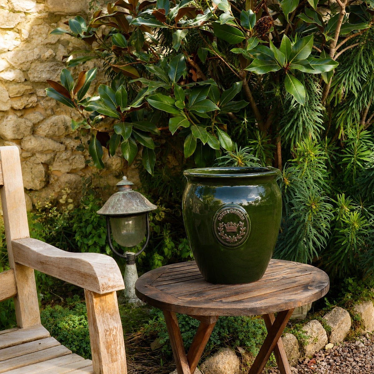 Der grün glasierte Blumentopf „eggpot“ (Höhe: 29–46 cm) steht auf einem runden Holztisch im Garten neben einem Holzstuhl und einer Vintage-Lampe, umgeben von üppigem Grün und einer Steinmauer als charmantem Hintergrund.