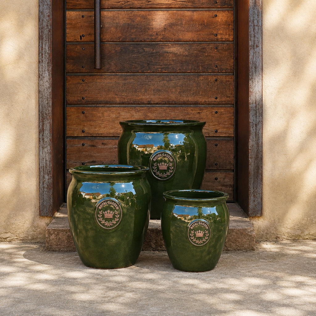 Drei grün glasierte Pflanztöpfe „eggpot“ (Höhen: 29–46 cm) stehen vor einer rustikalen Holztür mit Steinumrandung und werfen sanfte Schatten. Jeder glänzende Topf ist auf der Vorderseite mit einem dekorativen Emblem verziert.