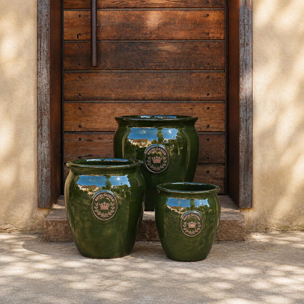 Drei grün glasierte Pflanztöpfe „eggpot“ (Höhen: 29–46 cm) stehen vor einer rustikalen Holztür mit Steinumrandung und werfen sanfte Schatten. Jeder glänzende Topf ist auf der Vorderseite mit einem dekorativen Emblem verziert.