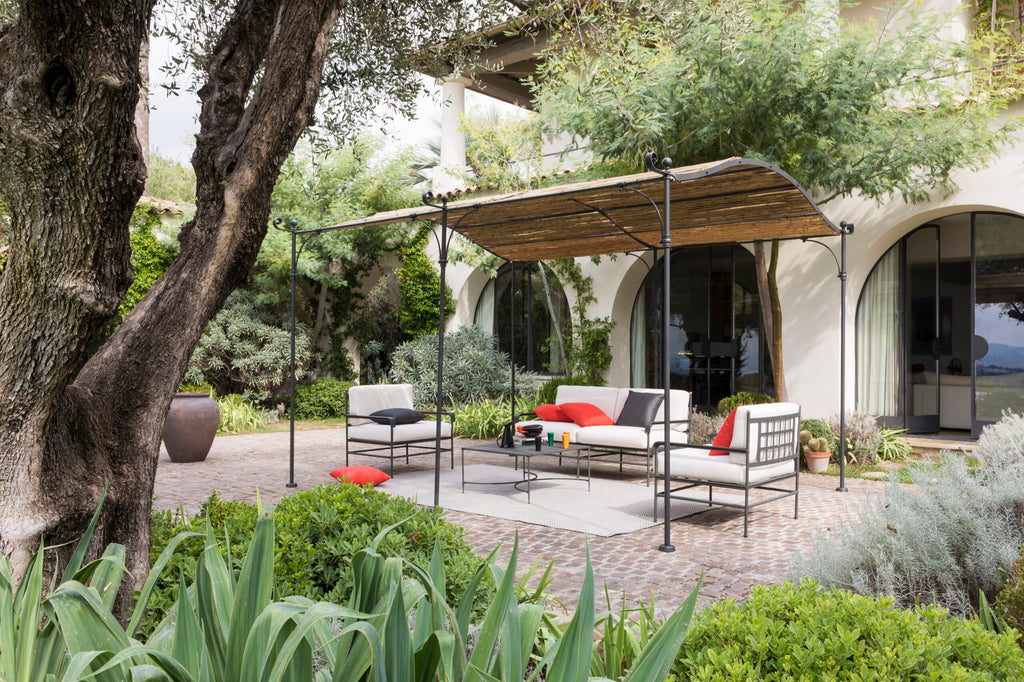 Eine ruhige Terrasse mit modernen Gartenmöbeln mit schwarzen Rahmen und weißen Kissen. Ein Sofa, zwei Stühle und ein Couchtisch stehen unter einer gewölbten Anbaupergola Solaire mit Holzschirm vor dem Hintergrund eines gewölbten Eingangs und üppigem Grün.