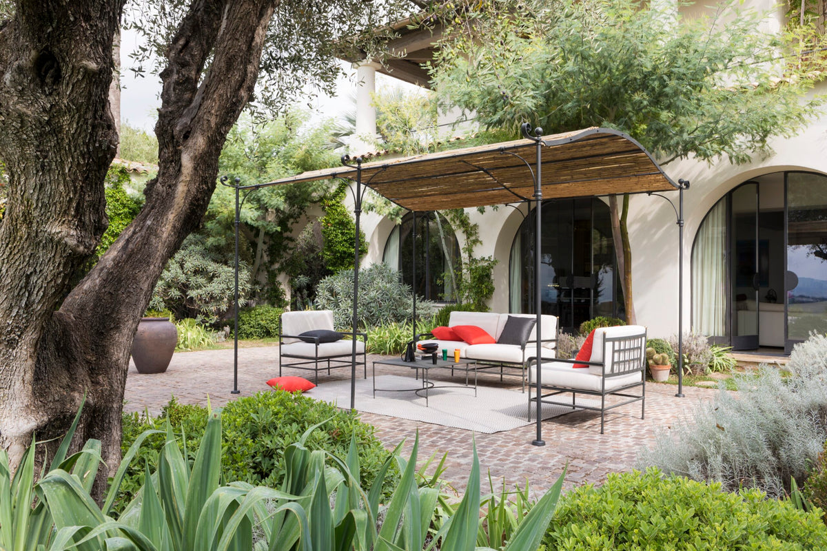 Eine ruhige Terrasse mit modernen Gartenmöbeln mit schwarzen Rahmen und weißen Kissen. Ein Sofa, zwei Stühle und ein Couchtisch stehen unter einer gewölbten Anbaupergola Solaire mit Holzschirm vor dem Hintergrund eines gewölbten Eingangs und üppigem Grün.