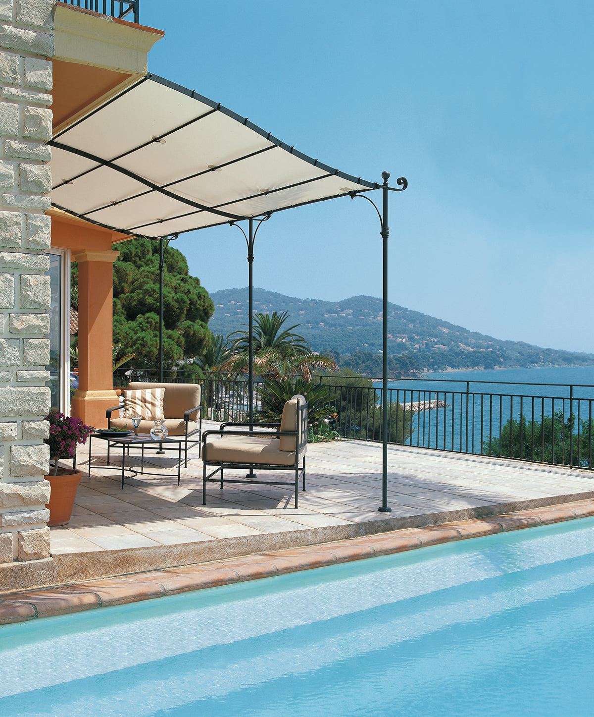 Eine luxuriöse Außenterrasse bietet gepolsterte Sitzgelegenheiten unter einem Dach aus Gebogene Anbaupergola Solaire mit Blick auf einen klaren blauen Pool. Angrenzend an ein Steinhaus bietet der Platz Ausblicke auf die fernen Berge und das Meer unter einem strahlend blauen Himmel. Üppiges Grün verstärkt die ruhige Atmosphäre.