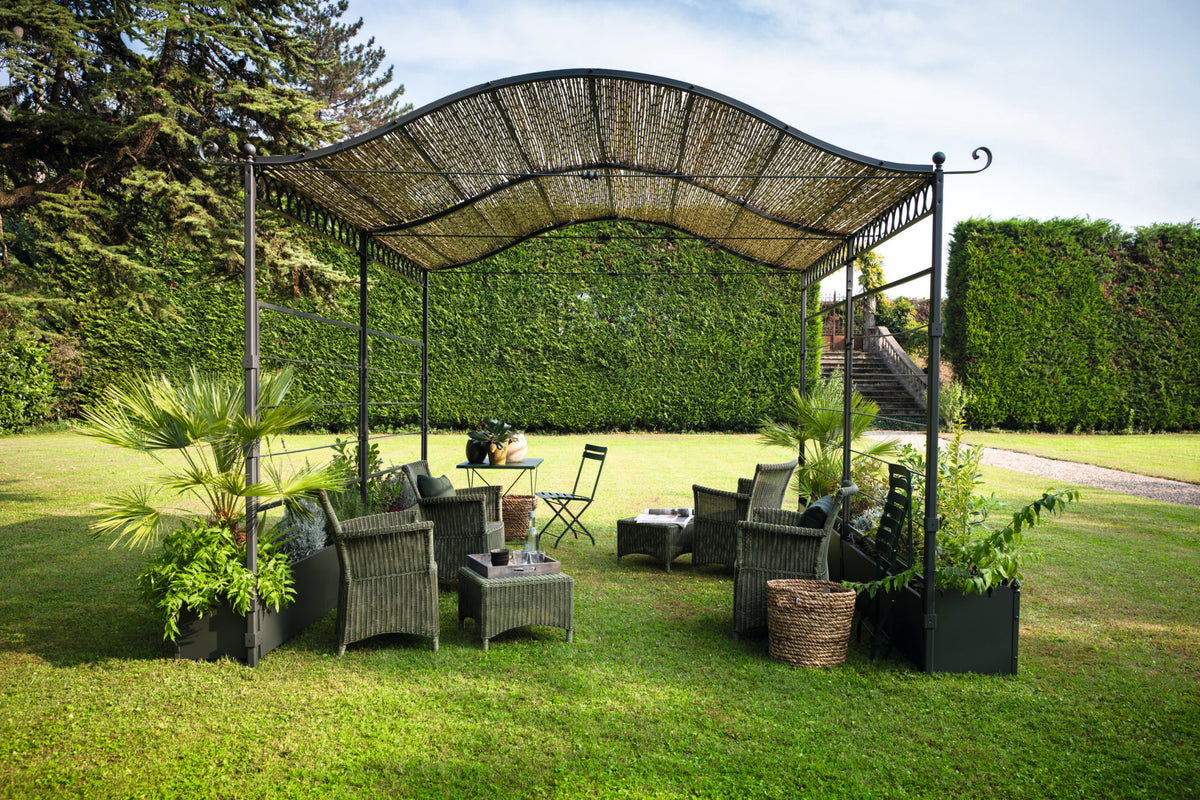 Die Gebogene Anbaupergola Solaire, ein grüner Außenpavillon mit geflochtenem, gewölbtem Dach, ist mit mehreren Korbstühlen und Tischen ausgestattet und von Topfpflanzen umgeben. Diese bezaubernde Pergola steht auf einem üppigen Rasen, eingerahmt von hohen grünen Hecken und ergänzt durch eine Treppe, die zu einem erhöhten Bereich im Hintergrund führt.