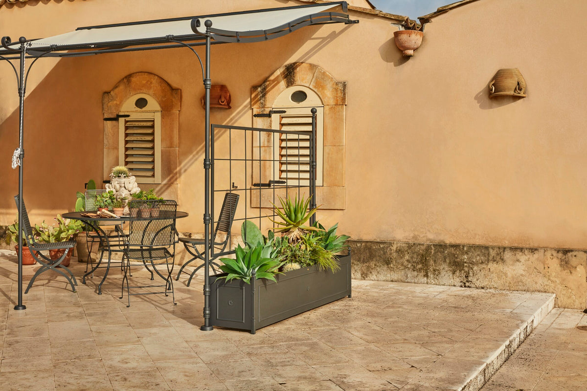 Eine sonnenbeschienene Außenterrasse mit einem Metalltisch und Stühlen befindet sich unter einer stilvollen, gebogenen Anbaupergola Solaire. Topfpflanzen schmücken den Bereich, begleitet von einer Gießkanne. Im Hintergrund befindet sich ein rustikales Steingebäude mit kleinen Fensterläden. Der Terrassenboden ist mit hellen Fliesen gepflastert und schafft so einen ruhigen Rückzugsort.