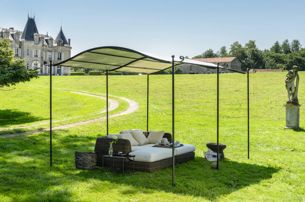 Ein ruhiger Sitzbereich im Freien mit gepolsterten Korbmöbeln und einer gebogenen Anbaupergola Solaire auf einem gepflegten Rasen. Im Hintergrund sind eine Statue und ein großes Steingebäude mit Türmchen zu sehen, die in natürliches Tageslicht getaucht sind und einen hellen und sonnigen Tag suggerieren.