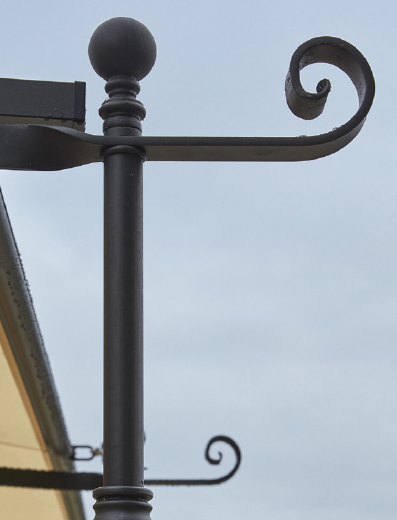 Nahaufnahme eines schwarzen schmiedeeisernen Wegweisers vor einem bewölkten Himmel. Der Pfosten hat eine dekorative Spitze mit runder Spitze und einem geschwungenen Ende, das die für das Design der Gebogenen Anbaupergola Solaire typische Eleganz ausstrahlt. Im Hintergrund sind zusätzliche Schnörkel zu sehen. Links ist eine Gebäudeecke zu sehen.