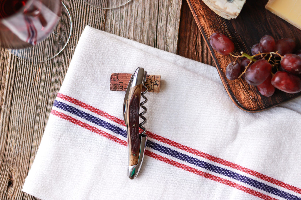 Die Forge De Laguiole Kellnermesser Sommelier | Fasseiche mit Kork ruht auf einem weißen Tuch mit roten und blauen Streifen, neben einem Holzbrett mit Trauben und Käse auf einem rustikalen Holztisch.
