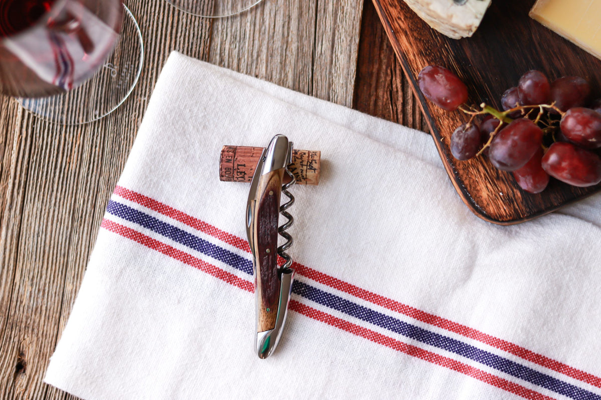 Die Forge De Laguiole Kellnermesser Sommelier | Fasseiche mit Kork ruht auf einem weißen Tuch mit roten und blauen Streifen, neben einem Holzbrett mit Trauben und Käse auf einem rustikalen Holztisch.