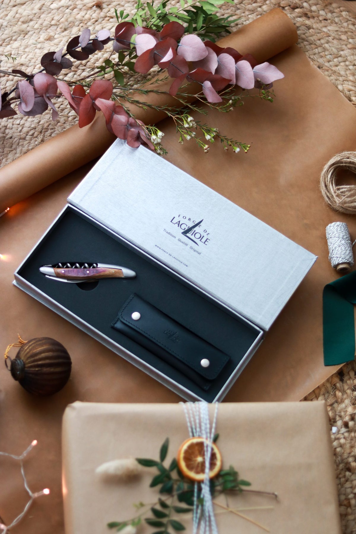 Ein Forge De Laguiole Kellnermesser Sommelier | Fasseiche, verpackt in einem Lederetui, ruht auf braunem Papier inmitten von getrockneten Blättern, Bindfäden, einer Kugel, grünem Band und einem eingepackten Geschenk mit getrockneten Zitrusfrüchten und Blättern.