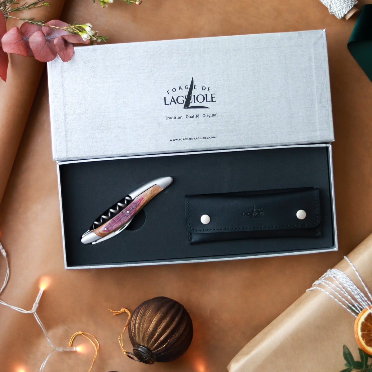 Das Forge De Laguiole Kellnermesser Sommelier | Fasseiche und sein schwarzes Lederetui werden in einer offenen grauen Schachtel mit der Aufschrift „Forge de Laguiole“ inmitten einer festlichen Weihnachtsdekoration präsentiert – ein elegantes Geschenk für Weinliebhaber.