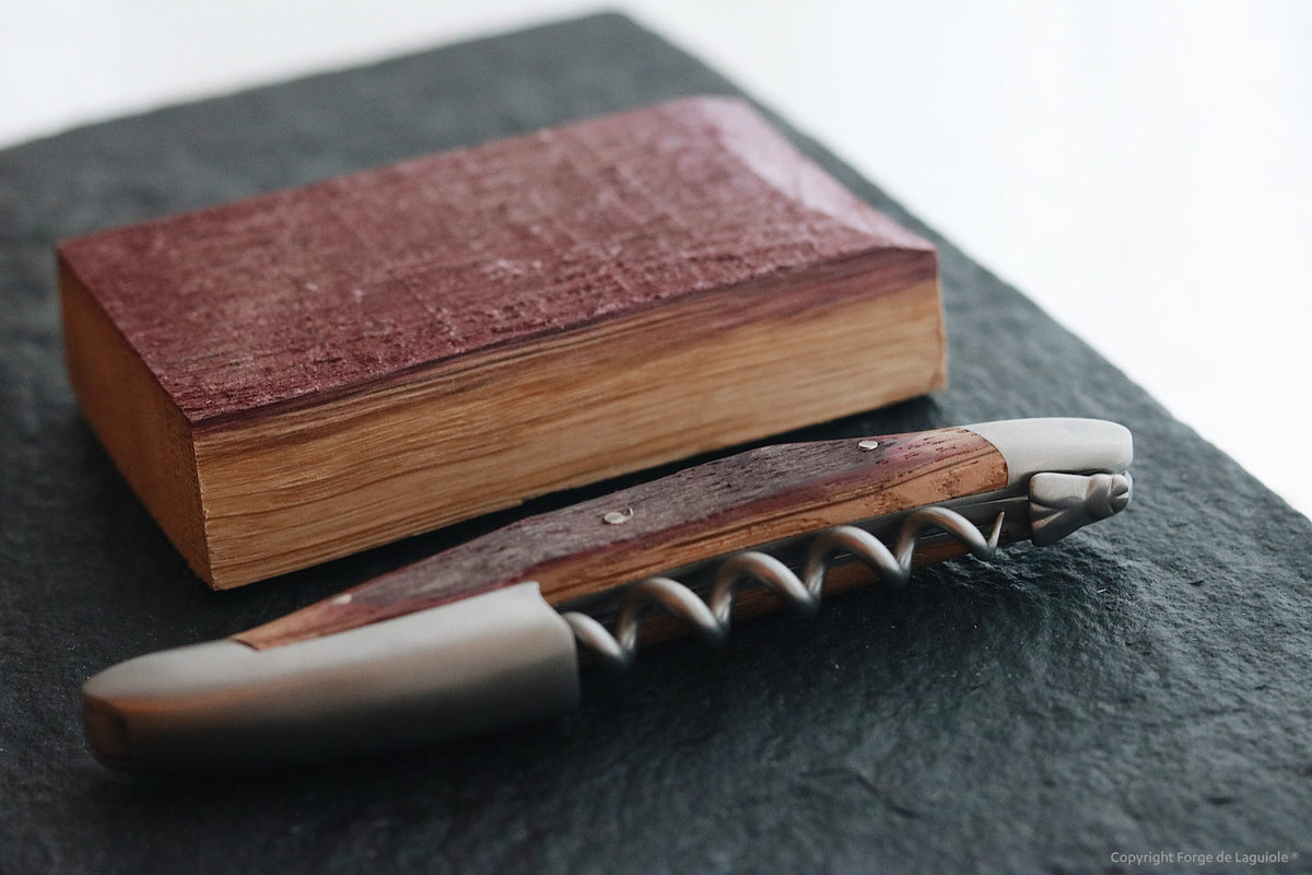 Ein dickes, geschlossenes Buch mit rötlich-braunem Einband liegt neben einer Forge De Laguiole Kellnermesser Sommelier | Fasseiche auf einer dunklen, strukturierten Oberfläche. Der Korkenzieher ist mit einer Spirale aus Edelstahl und dekorativen Akzenten versehen.