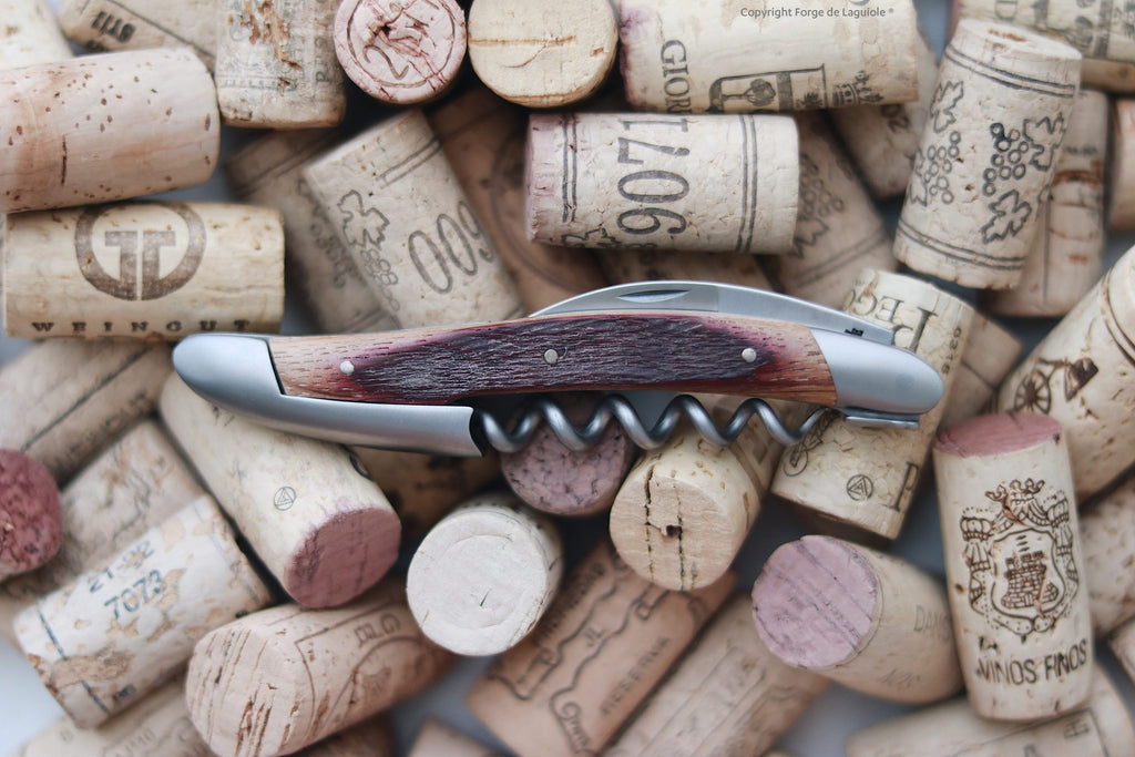 Die Kellnermesser Sommelier | Fasseiche von Forge De Laguiole mit einem Holzgriff aus Fasseiche ruht auf einem Haufen gebrauchter Weinkorken, die teilweise rot gefärbt sind und einzigartige Winzerlogos tragen.