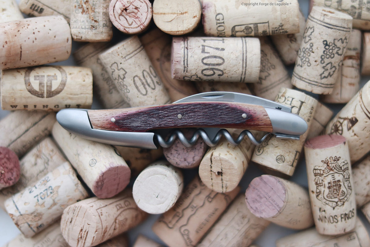 Die Kellnermesser Sommelier | Fasseiche von Forge De Laguiole mit einem Holzgriff aus Fasseiche ruht auf einem Haufen gebrauchter Weinkorken, die teilweise rot gefärbt sind und einzigartige Winzerlogos tragen.