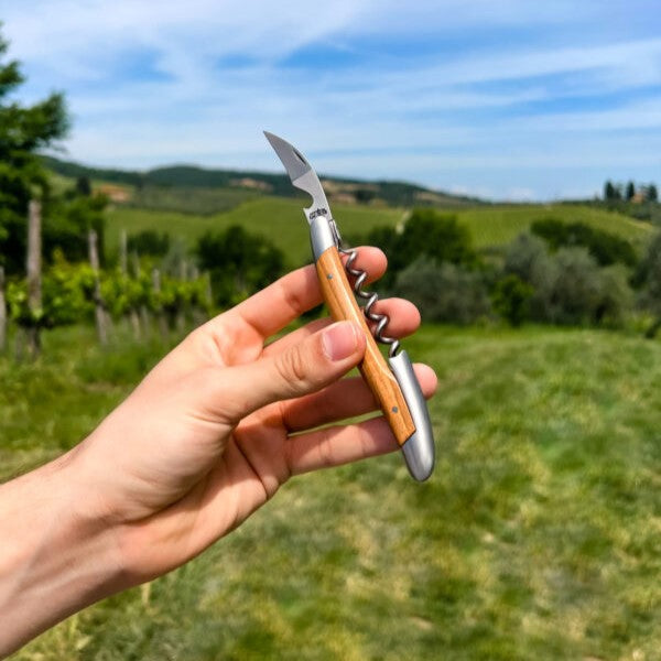 Das Kellnermesser Sommelier | Olive von Forge De Laguiole mit seinem Holzgriff wird gekonnt vor der Kulisse üppig grüner Weinberge unter einem klaren blauen Himmel gehalten und spiegelt die typische Präzision eines Sommeliers wider.