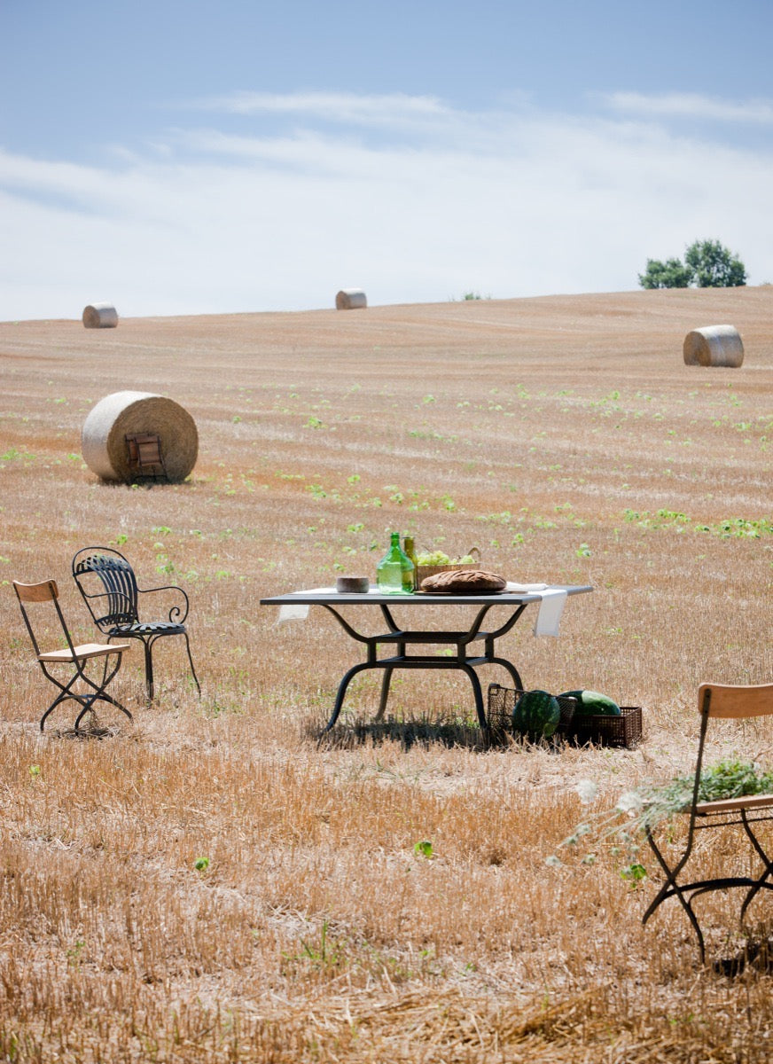 Ein ruhiger Essbereich im Freien auf einem sonnenbeschienenen Feld mit verstreuten Heuballen. Die Szene zeigt einen weißen Tisch mit Essen und Getränken, umgeben von drei Sesseln Toscana aus Eisen in Graphitgrau. Das weite Feld und der klare Himmel schaffen eine friedliche, rustikale Atmosphäre im toskanischen Stil.