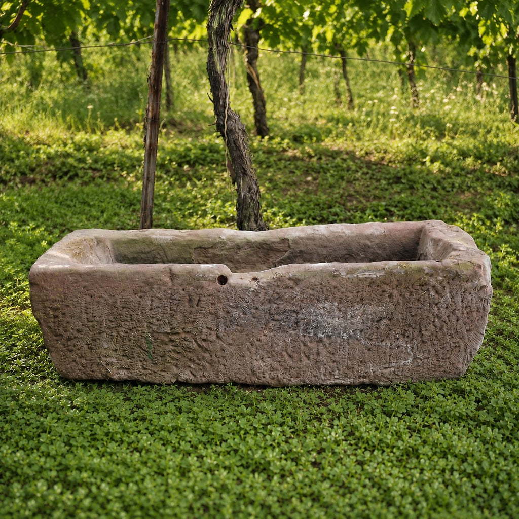 Die Sandstein Vorlage - ohne Loch (Kopie), ein großer rechteckiger Steintrog, steht auf grünem Gras inmitten von üppigen Weinreben und Bäumen in einem sonnigen Weinberg.