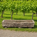 Ein altertümlicher Futtertrog aus Sandstein (152 x 39 x 25) steht auf Blöcken vor einer Reihe von Weinreben in einem Weinberg, im Vordergrund sind grünes Gras und Kies zu sehen.