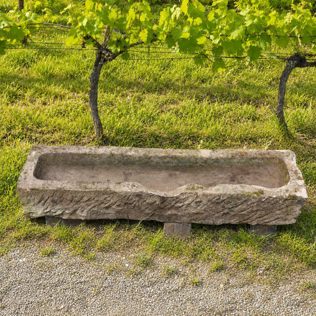 Ein altertümlicher Futtertrog aus Sandstein (152 x 39 x 25) steht auf Gras und Kies vor einer Reihe von Weinreben mit grünen Blättern.
