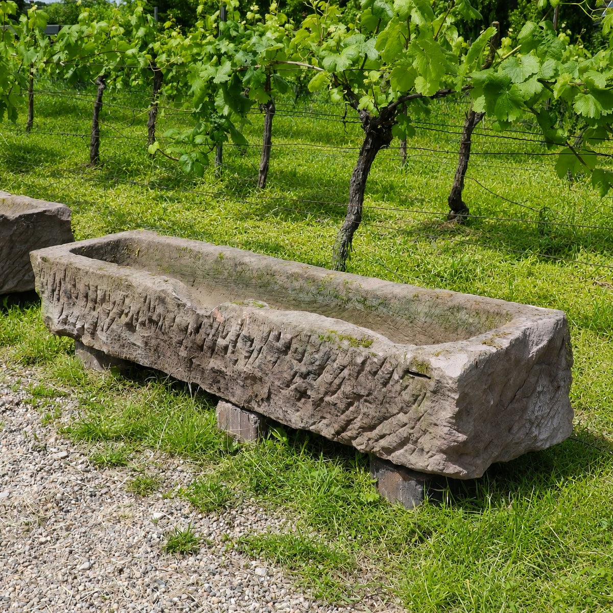 Ein „altertümlicher Futtertrog aus Sandstein, 152 x 39 x 25“ steht auf einer Rasenfläche neben einem Schotterweg, eingerahmt von Weinreben und üppigem Grün im Hintergrund.