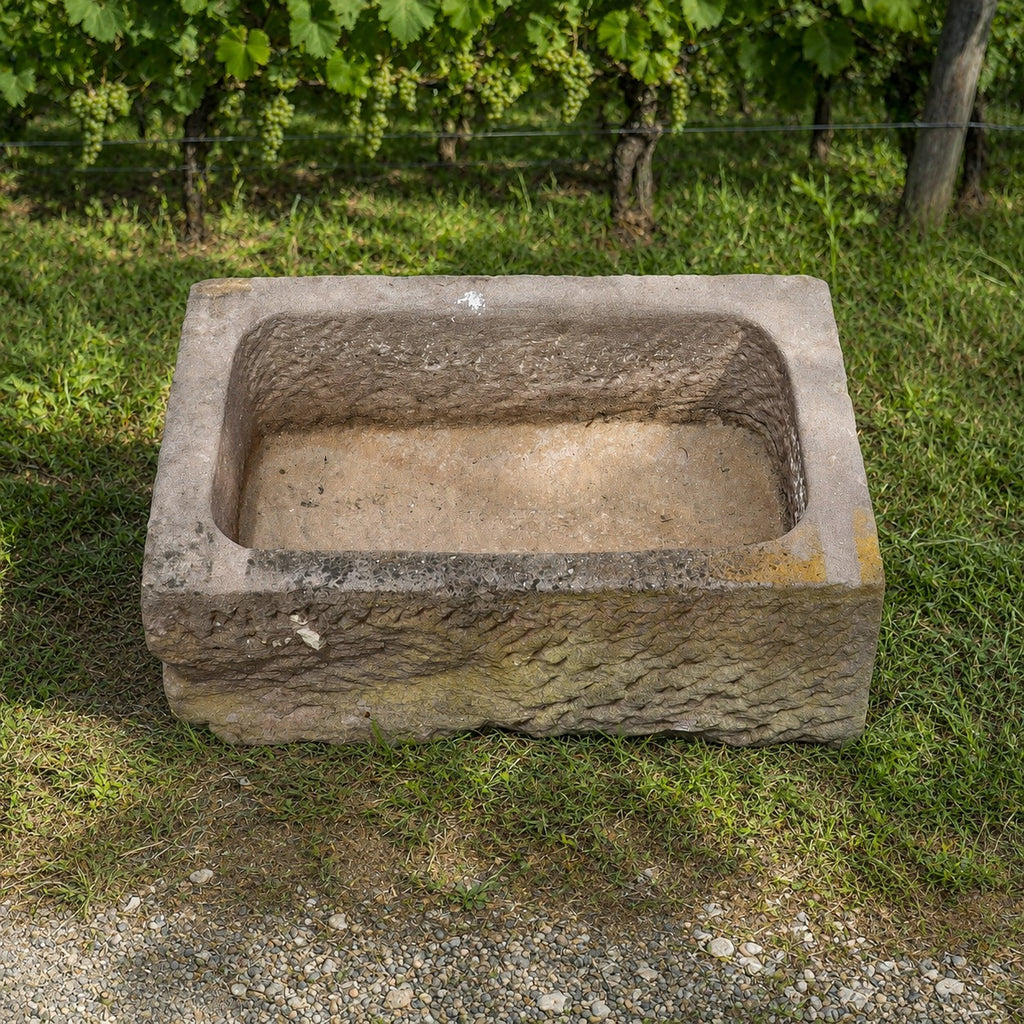 Ein Natursteinbecken aus Sandstein (78 x 53 x 29), das eine rechteckige Form hat und an traditionelle Steintrog erinnert, steht auf einer Rasenfläche neben einem Kiesweg, während im Hintergrund ein Weinberg und üppig grüne Weinreben zu sehen sind.