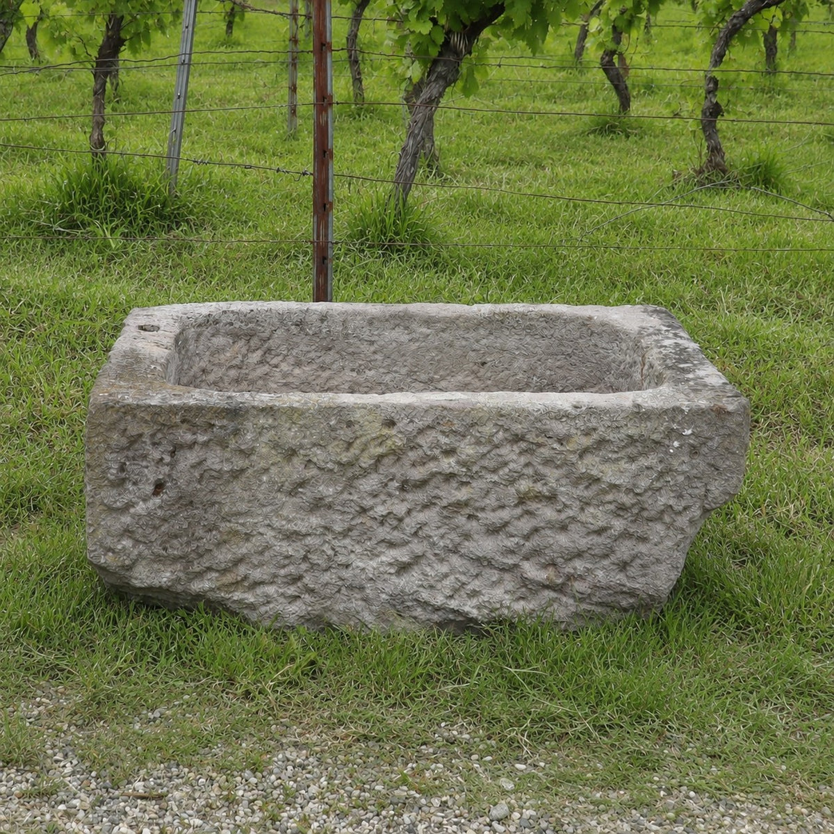 Ein Sandsteintrog mit den Maßen 84 x 55 x 32, der sich durch ein rustikales, rechteckiges Design auszeichnet, steht auf grünem Gras, im Hintergrund sind ein Weinberg und ein Drahtzaun zu sehen.