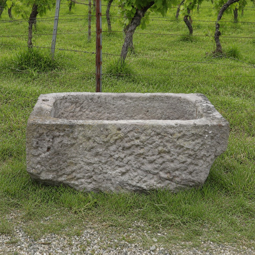 Ein Sandsteintrog mit den Maßen 84 x 55 x 32, der sich durch ein rustikales, rechteckiges Design auszeichnet, steht auf grünem Gras, im Hintergrund sind ein Weinberg und ein Drahtzaun zu sehen.