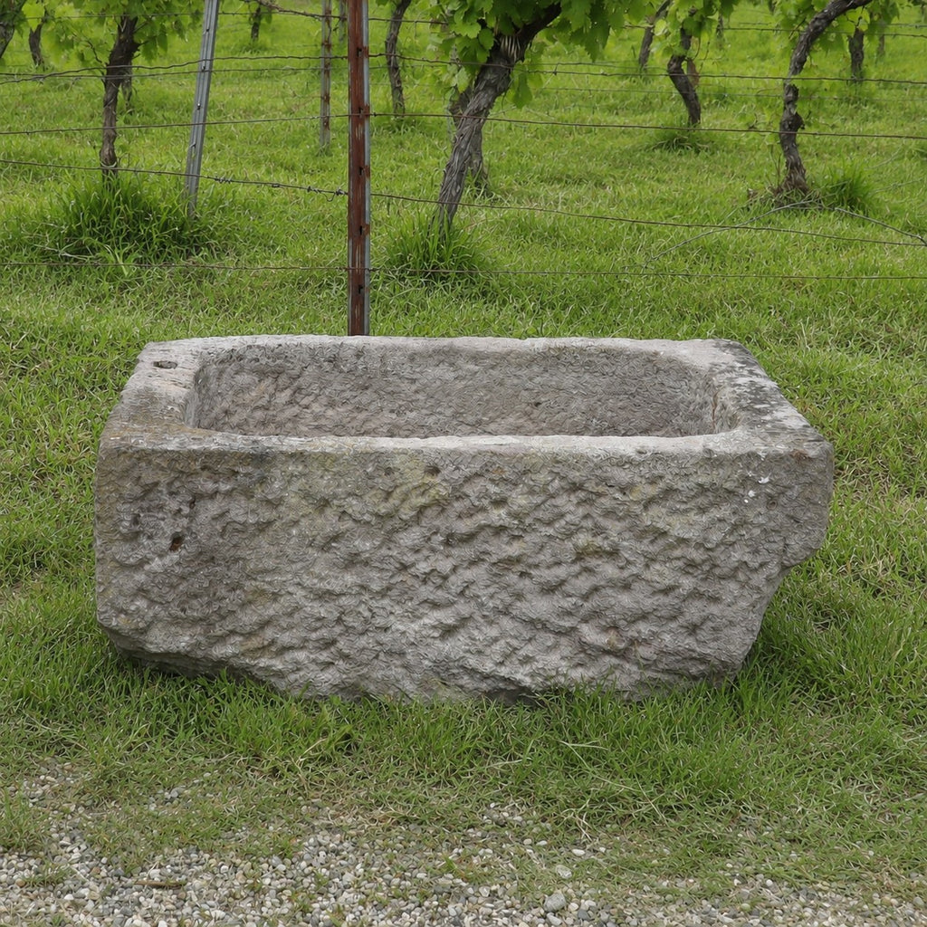 Ein Sandsteintrog mit den Maßen 84 x 55 x 32, der sich durch ein rustikales, rechteckiges Design auszeichnet, steht auf grünem Gras, im Hintergrund sind ein Weinberg und ein Drahtzaun zu sehen.