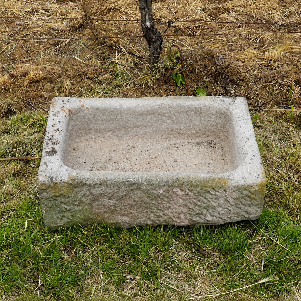 Ein alter Sandstein-Trog (84 × 55 × 32, Kopie), rechteckig und verwittert, steht auf Gras und trockenem Stroh; dahinter sind Erde und eine kleine Pflanze zu sehen.