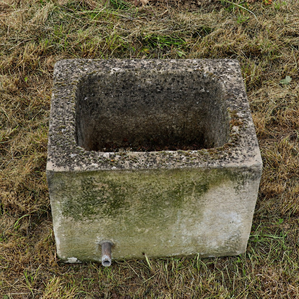 Ein verwitterter, rechteckiger Brunnentrog (55 × 43 × 40) aus altem Sandstein, dessen Oberfläche mit Moos und Algen bewachsen ist, steht auf trockenem Gras. An einer Seite befindet sich ein kleiner Metallauslauf, während die offene Oberseite den hohlen Innenraum freigibt.