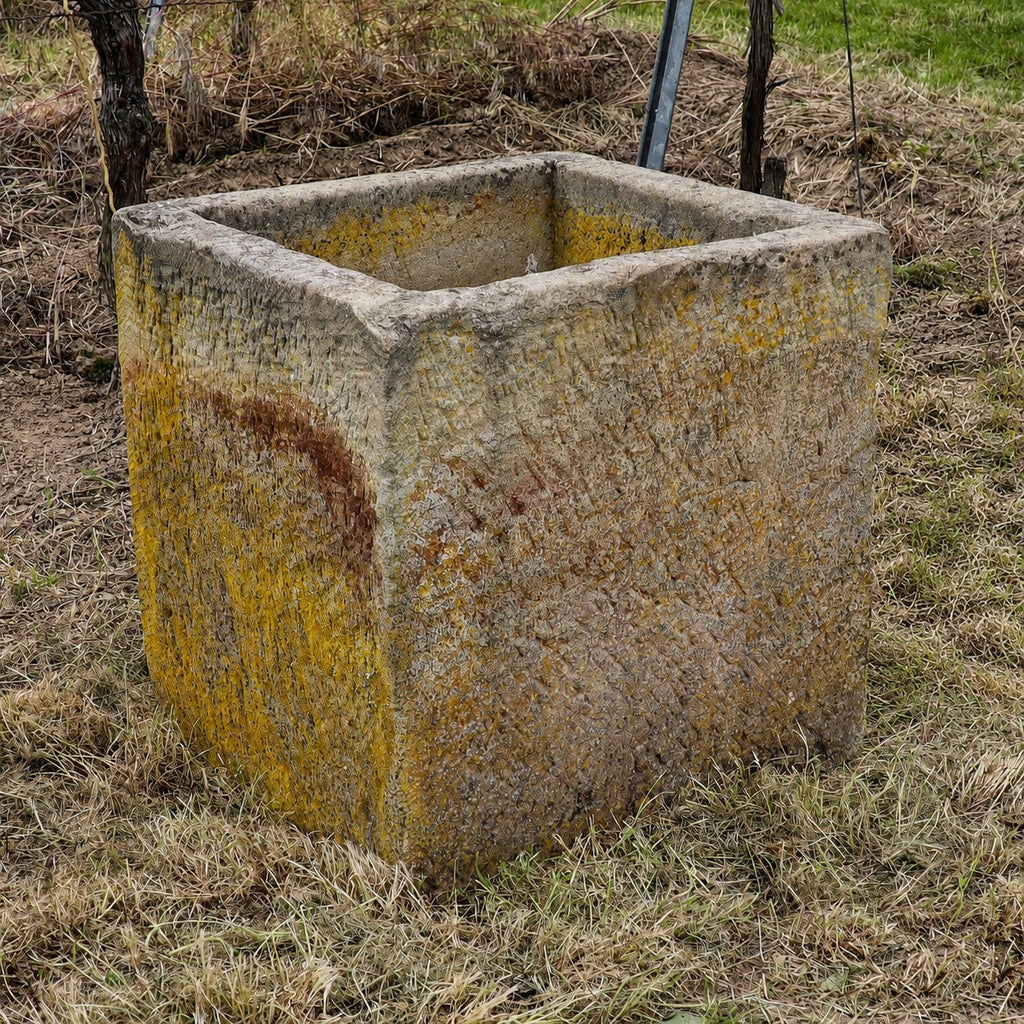 Ein historischer Sandsteinkübel (62 x 59 x 60) mit gelben und braunen Flecken steht auf trockenem Gras in der Nähe eines Feldes.